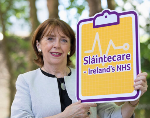 Róisín Shortall TD and member of the Social Democrats is standing holding a board designed to look like a clipboard. The board has a yellow background and purple text that says Sláintecare - Ireland's NHS. The woman is smiling and wearing a black top and light coloured jacket.